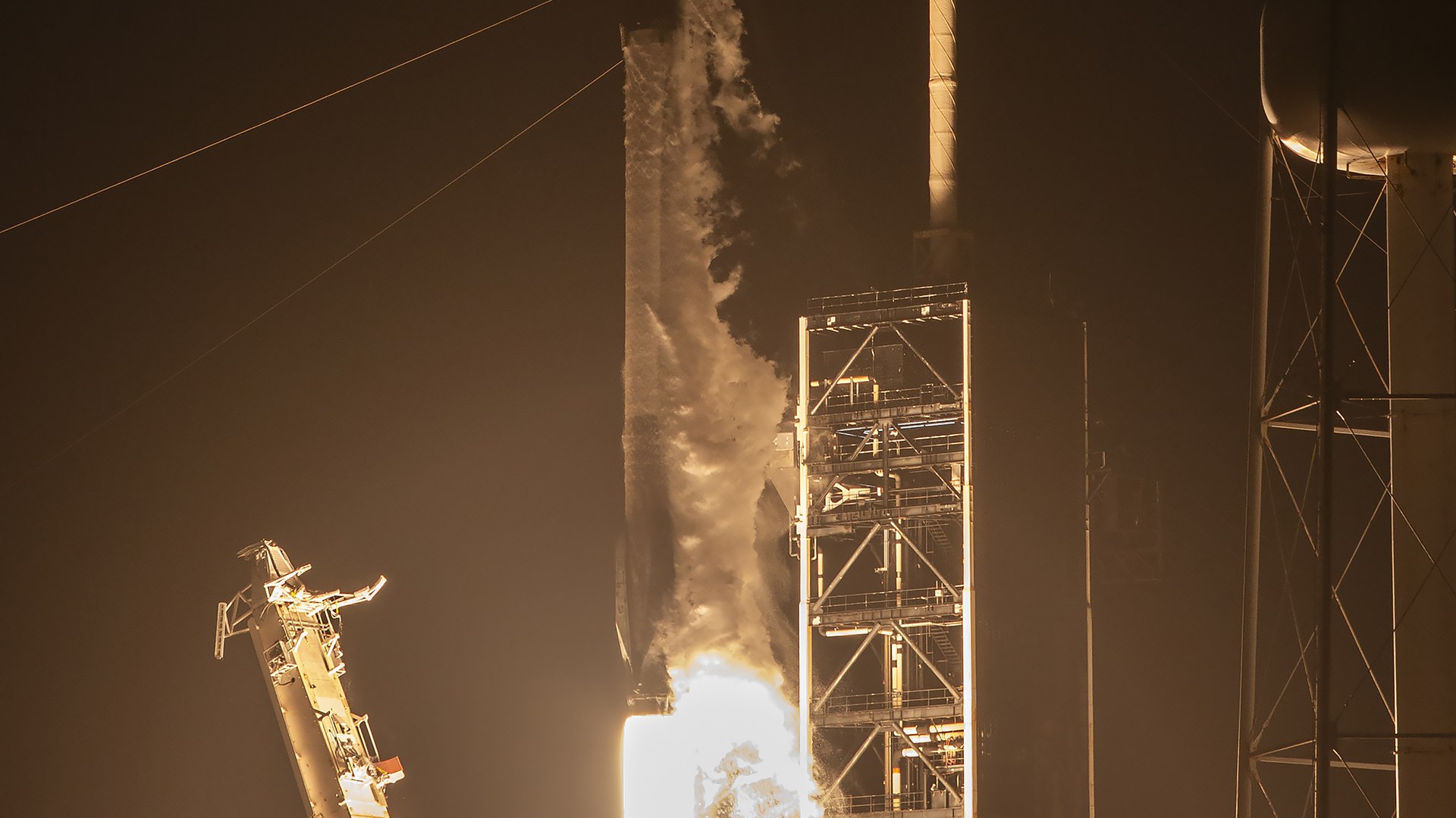 Foguete Falcon 9 da SpaceX decolando de madrugada ao lado da torre de lançamento, com o fogo dos motores iluminando a fumaça e a estrutura metálica no escuro