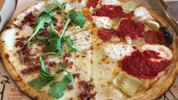 Pizza redonda do Blaze Pizza numa caixa de papelão, dividida em sabores: metade com rúcula, bacon e queijo branco, metade com molho de tomate, pepperoni e alcachofra