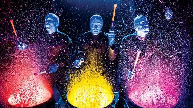 Três atores de pele pintada de azul tocam tambores com baquetas no palco escuro, e a tinta vermelha, amarela e rosa espirra para cima de cada tambor iluminado