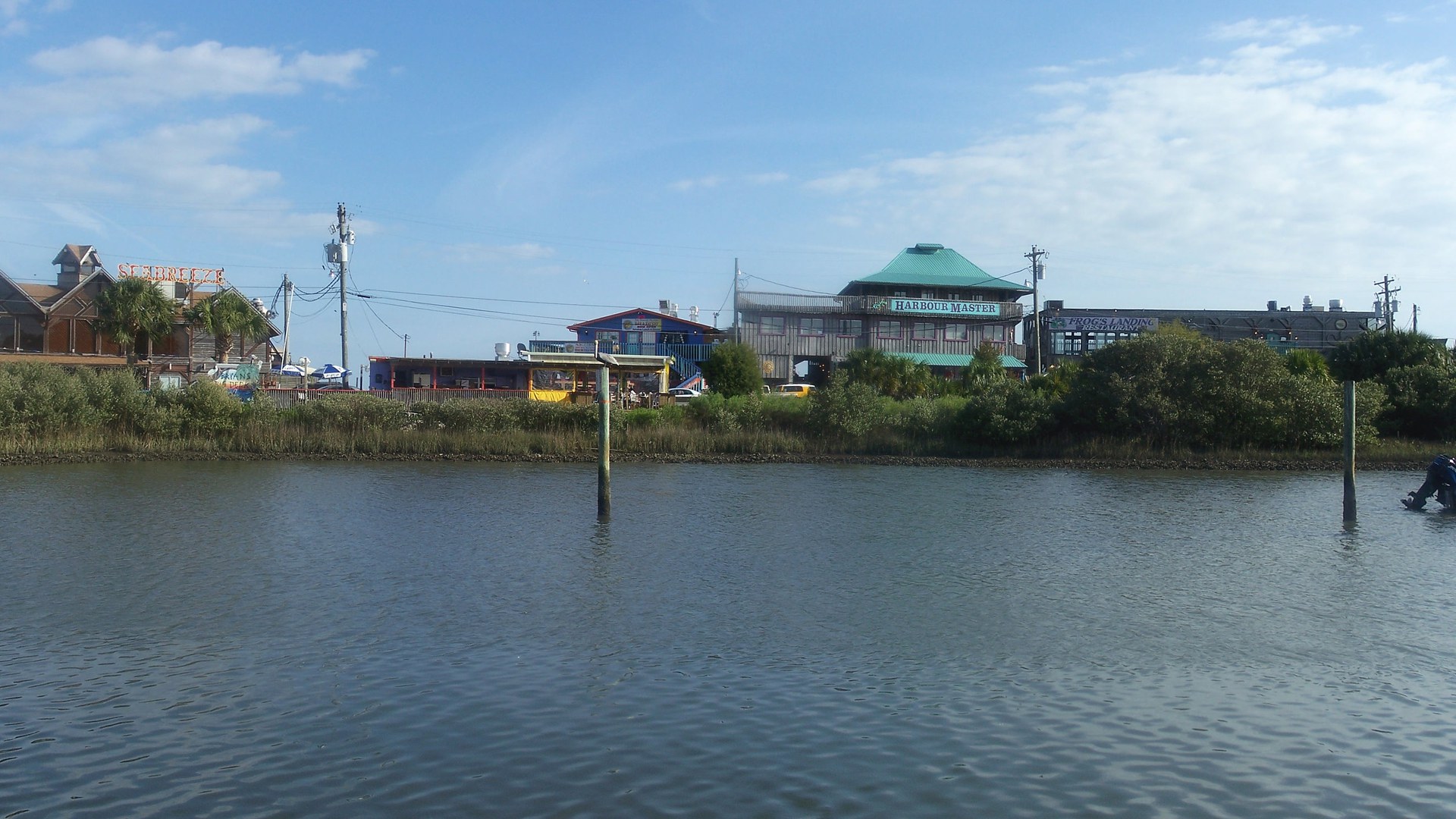 A Dock Street de Cedar Key vista da água: restaurantes e lojas de madeira coloridos, um prédio de telhado verde com a placa Harbour Master, mangue na margem e água calma em primeiro plano, sob céu azul