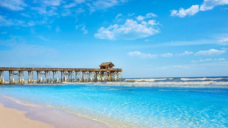 Píer de madeira com um quiosque coberto na ponta avançando sobre o mar azul-turquesa, com areia clara e espuma das ondas em primeiro plano; foto do acervo, de cores realçadas, que parece mostrar o Cocoa Beach Pier