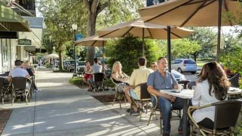 Pessoas sentadas em mesas de café sob guarda-sóis na calçada arborizada de uma rua comercial, com carros estacionados e árvores ao fundo
