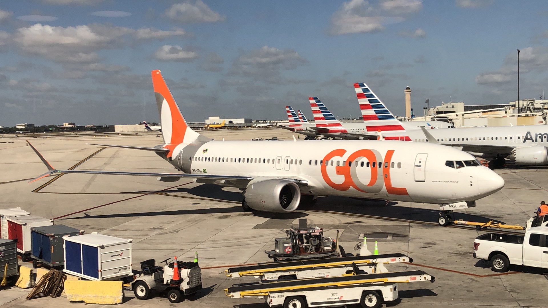 Boeing 737 MAX 8 branco e laranja da Gol estacionado no pátio do aeroporto de Miami, com carrinhos de bagagem em primeiro plano e caudas de aviões da American Airlines ao fundo