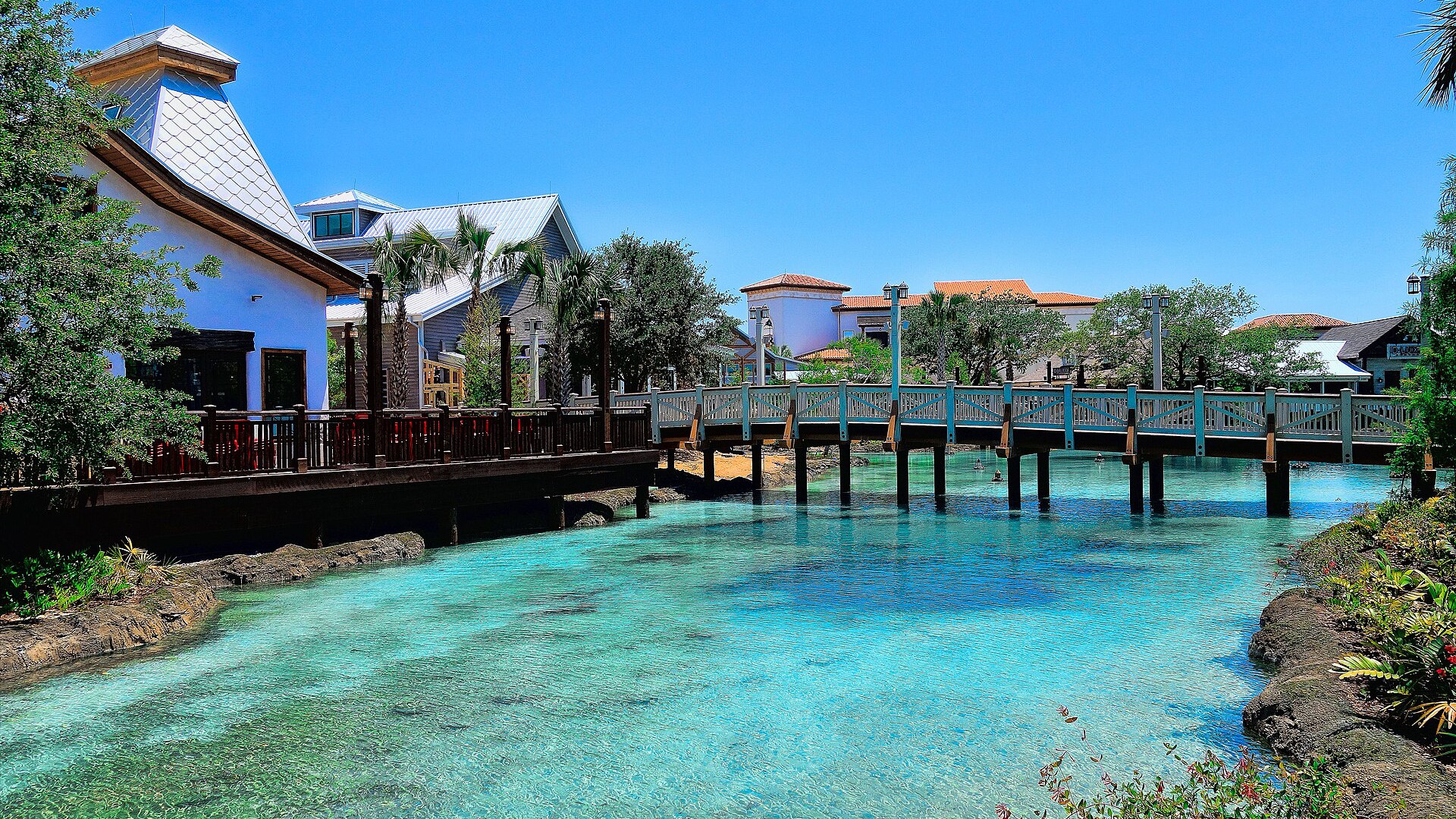 Ponte de madeira sobre o canal de água azul-turquesa da Disney Springs, com prédios de telhado claro e palmeiras dos dois lados, num dia de céu limpo