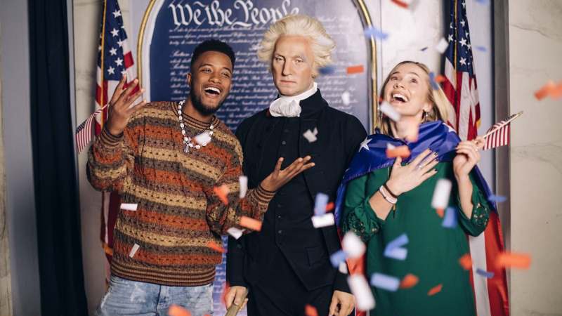 Dois visitantes rindo ao lado da figura de cera de um presidente americano de peruca branca e casaca preta, diante de um painel com «We the People» e bandeiras dos Estados Unidos, sob uma chuva de confete