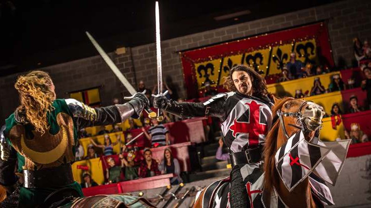 Dois cavaleiros cruzam espadas na arena do Medieval Times, um de verde e dourado e outro de preto e branco com cruz vermelha, montado num cavalo com a mesma armadura, com o público na arquibancada ao fundo