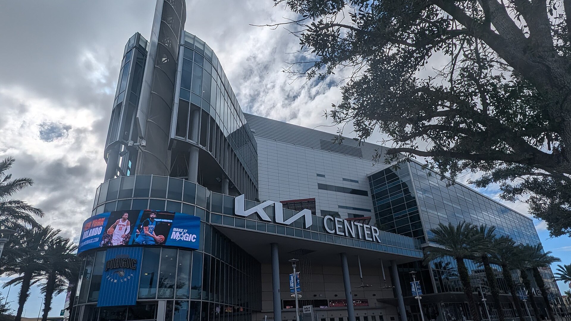 Fachada do Kia Center, no centro de Orlando, com o letreiro da Kia, a torre de vidro e o painel de LED do Orlando Magic sobre a loja do time, sob céu nublado