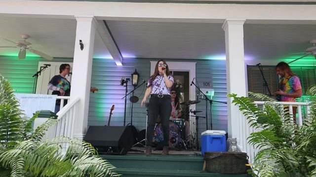 Uma banda toca na varanda de uma casa de madeira verde-clara: a cantora ao microfone na frente, guitarristas dos lados e a bateria ao fundo, com samambaias na escada