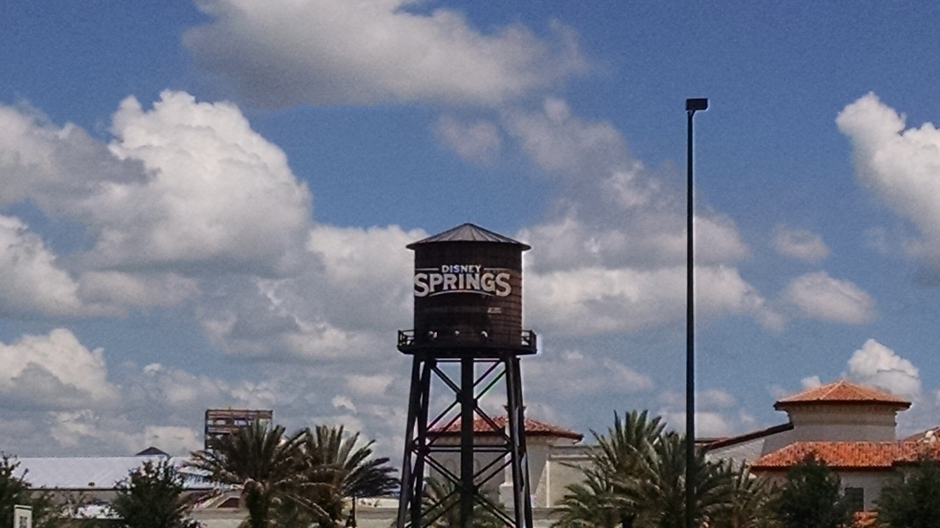 Caixa d'água de madeira com o letreiro «Disney Springs» sob céu azul com nuvens, com palmeiras e telhados ao fundo.