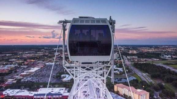 Cabine da roda-gigante no alto da estrutura branca, ao entardecer, com a International Drive, estacionamentos e hotéis lá embaixo e o céu rosado no horizonte