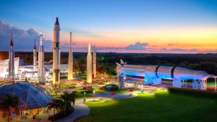 Jardim de foguetes do Kennedy Space Center Visitor Complex ao entardecer, com foguetes de pé iluminados, um foguete deitado iluminado em azul e uma cúpula de vidro no primeiro plano, em foto antiga do acervo que mostra o complexo, e não a cidade de Titusville