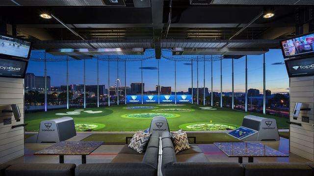 Baia do Topgolf ao anoitecer, com sofá, mesas, telas de TV e as máquinas de bola, de frente para o campo de grama iluminado com alvos coloridos, a rede alta em volta e, ao fundo, prédios e uma roda-gigante
